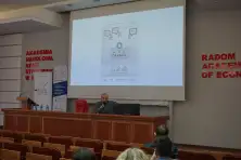 Prelegent przy stole prezydialnym, na ekranie slajd prezentacji